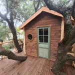 Cabane en bois perchée, entourée darbres, avec une porte verte et un balcon en bois.
