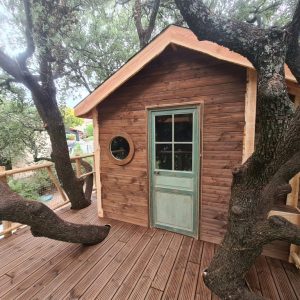 Cabane en bois perchée, entourée darbres, avec une porte verte et un balcon en bois.