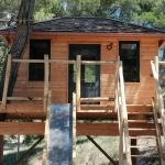 Cabane en bois perchée dans les arbres, avec un escalier en bois et de grandes fenêtres.