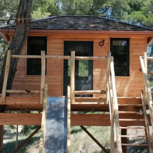 Cabane en bois perchée dans les arbres, avec un escalier en bois et de grandes fenêtres.