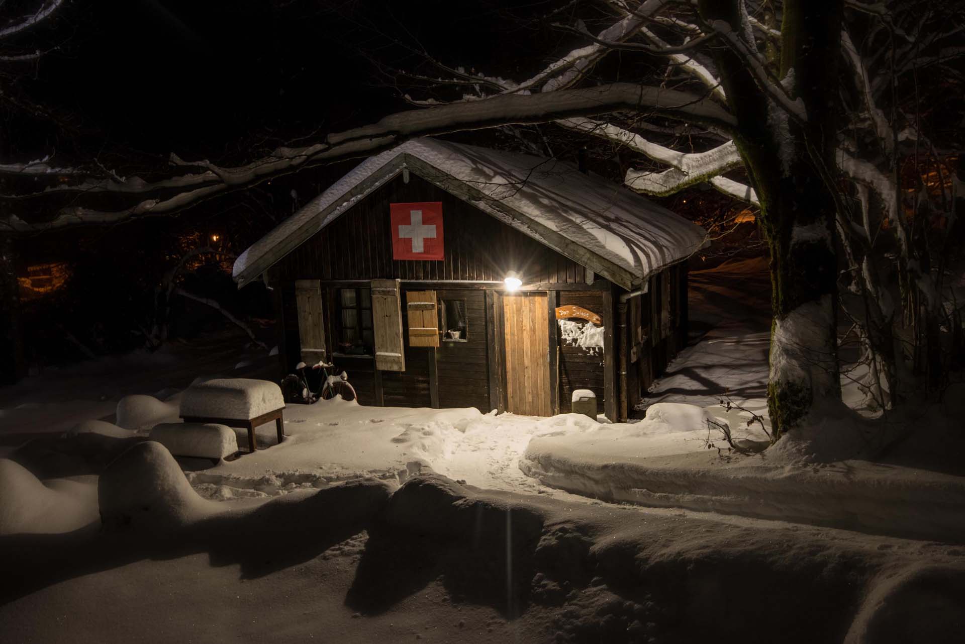 location-insolite-location-hiver-boldair Cabane suisse en bois, illuminée, entourée de neige et de branches enneigées.