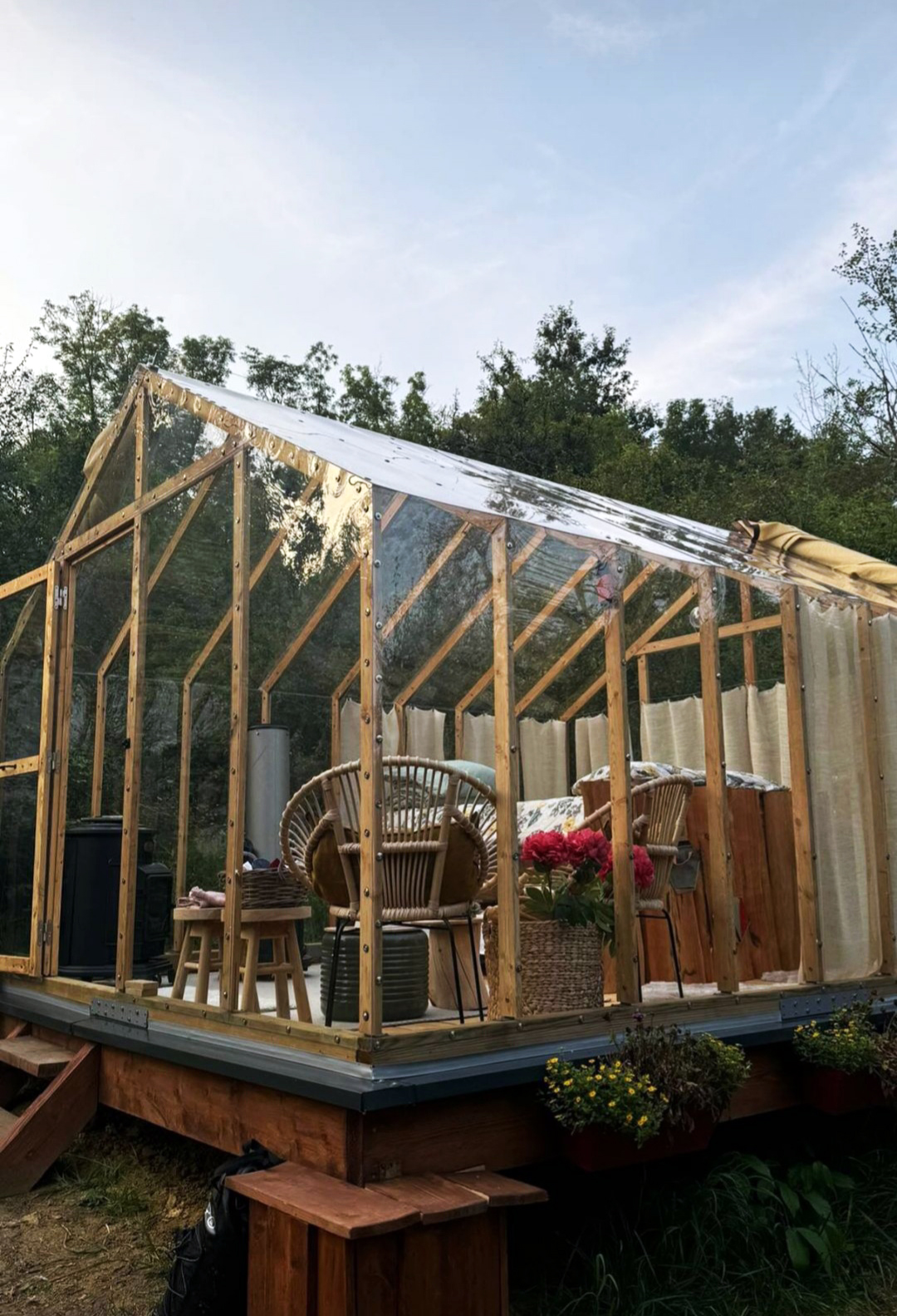 1725884271555 (1) Cabane en verre dans la nature, avec mobilier en rotin et fleurs colorées.