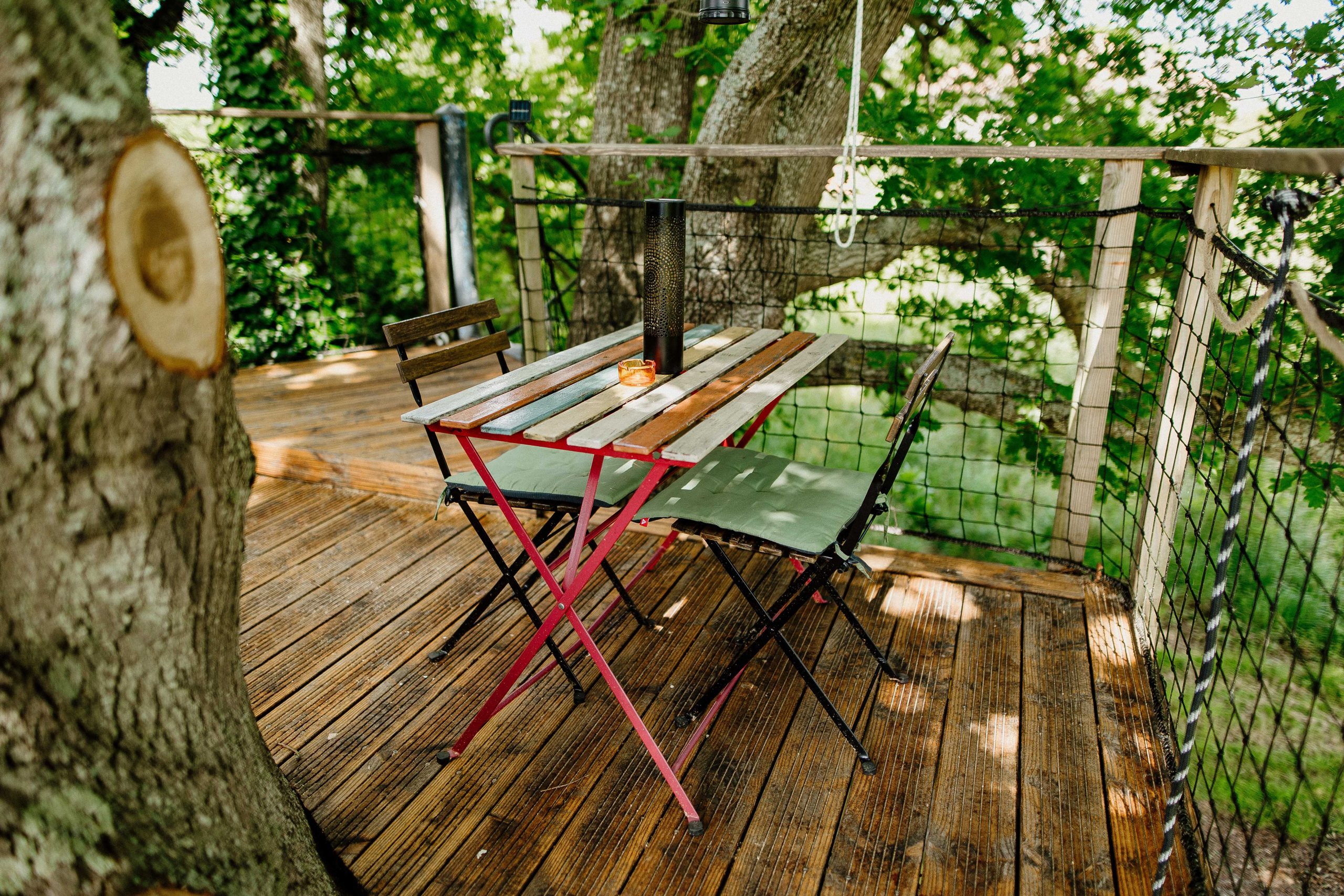 Cabane dans les arbres avec une terrasse en bois, table et chaises en métal, entourée de verdure.