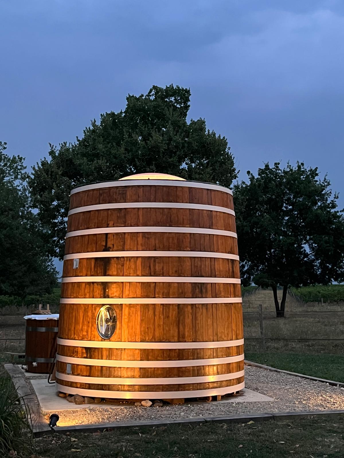 964b36d4-8fa5-420c-9f8d-fe5d8953f1fb Hébergement insolite : un baril en bois éclairé, entouré d'arbres, sous un ciel nuageux.