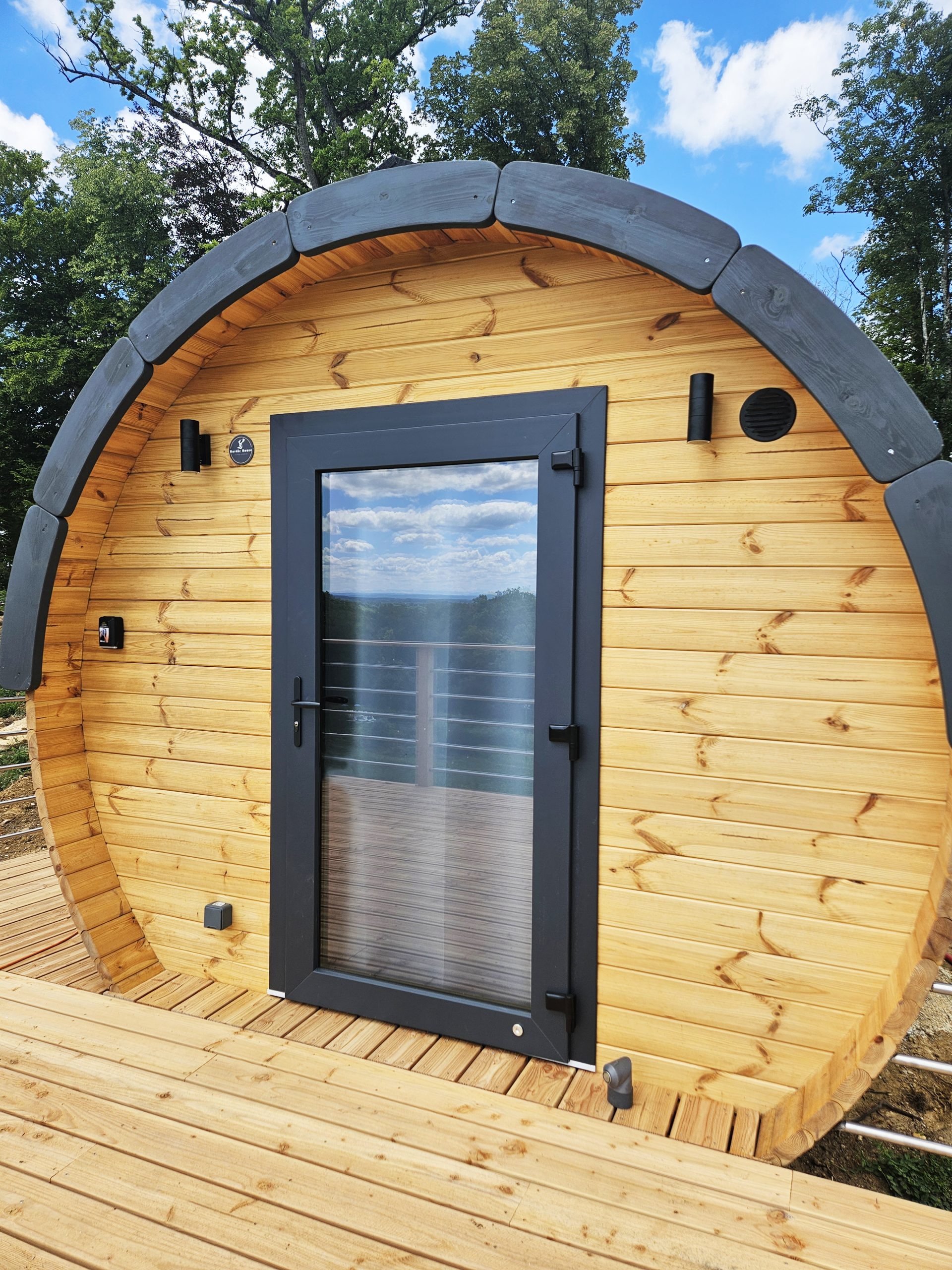 barrel_bouton_or Cabane en bois en forme de tonneau, avec une porte vitrée et un balcon en bois.