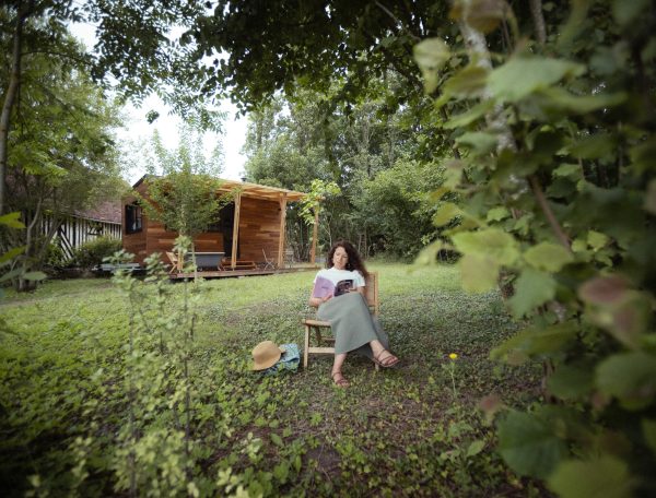 Cabane en bois entourée de verdure, femme lisant paisiblement à lextérieur.