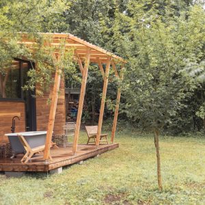Cabane en bois avec terrasse ensoleillée et baignoire extérieure, entourée de verdure.