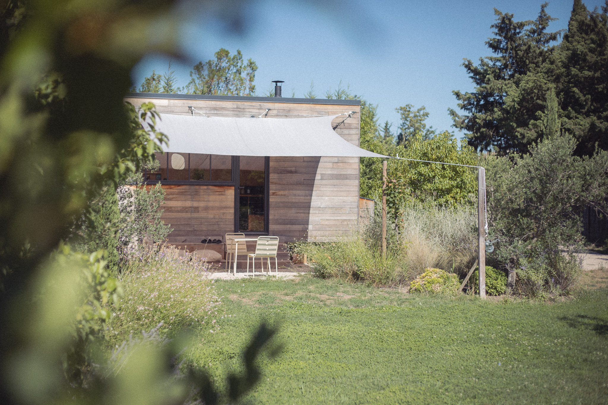 1L6A7193 Cabane en bois avec terrasse ombragée, entourée de verdure et d'un jardin fleuri.