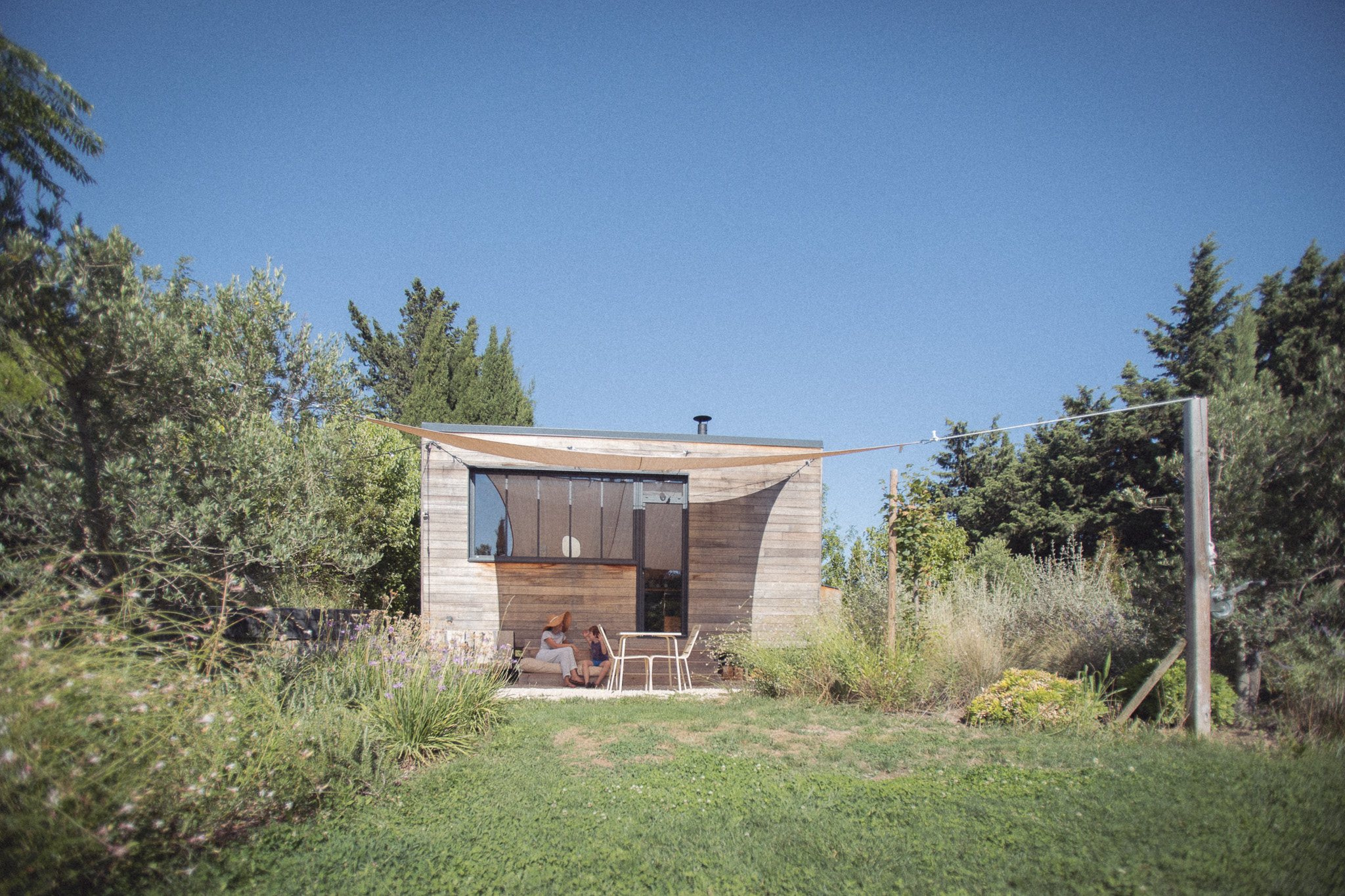1L6A7236 Cabane moderne en bois, entourée de verdure, avec une terrasse ombragée et un ciel bleu.