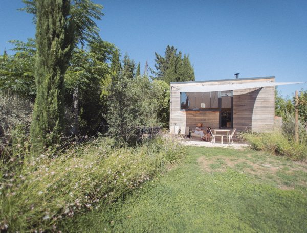 Cabane en bois moderne entourée de verdure, avec une terrasse et un ciel bleu.