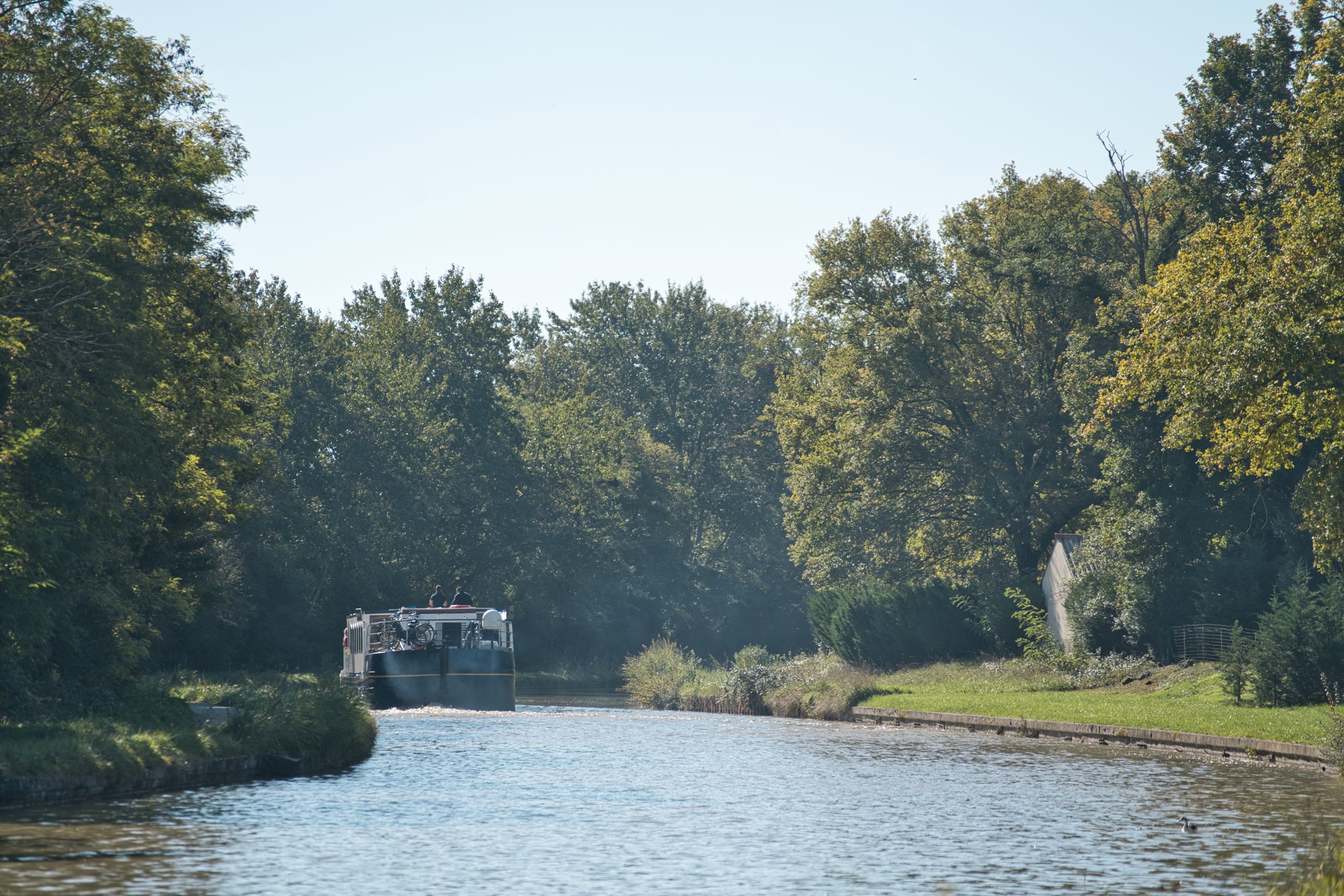 Péniche sur un canal, entourée darbres verdoyants sous un ciel bleu.