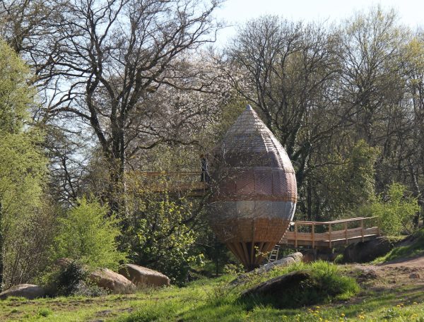Cabane dans les arbres en forme de goutte, entourée de verdure et de rochers.