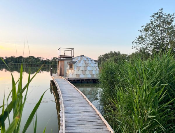 Dôme flottant sur un lac, accessible par une passerelle en bois, entouré de verdure.