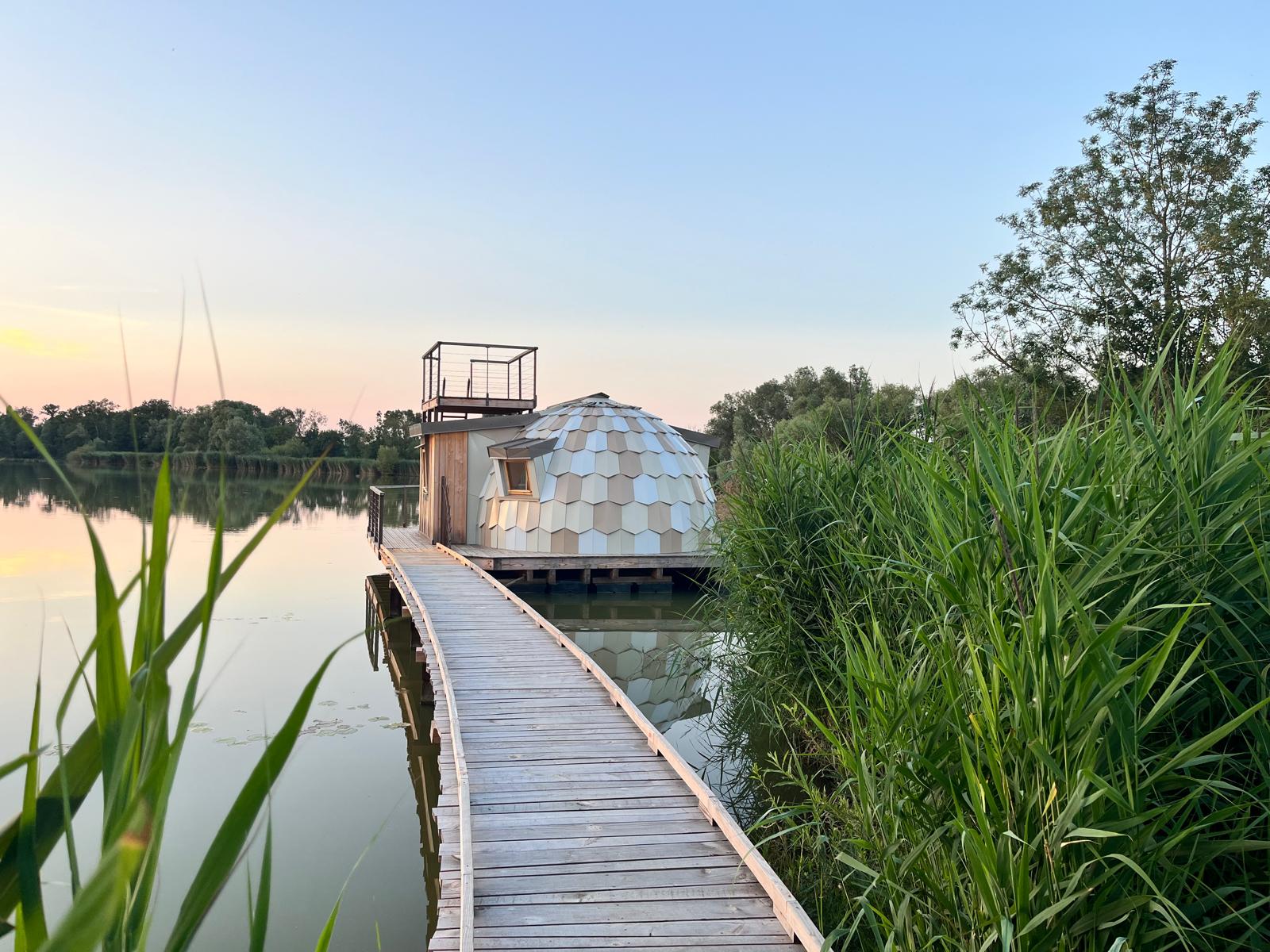 Dôme flottant sur un lac, accessible par une passerelle en bois, entouré de verdure.