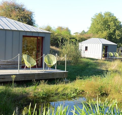 Hébergement en cabanes modernes avec chaises vertes au bord dun étang paisible.