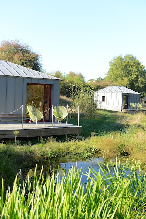 Hébergement en cabanes modernes avec chaises vertes au bord dun étang paisible.