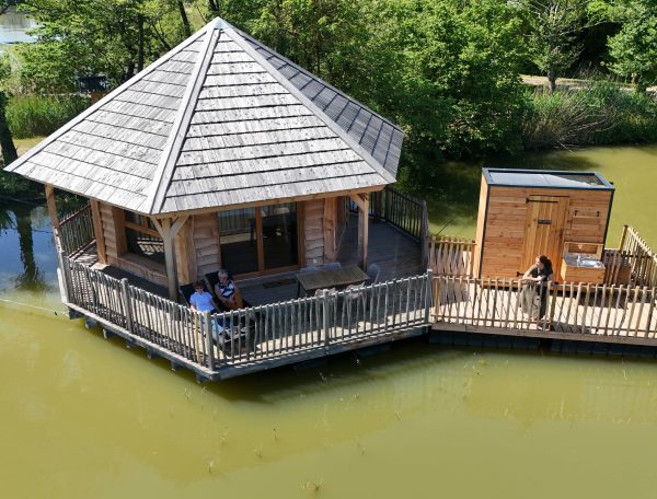 Cabane sur leau avec terrasse en bois, entourée de verdure et dun étang paisible.