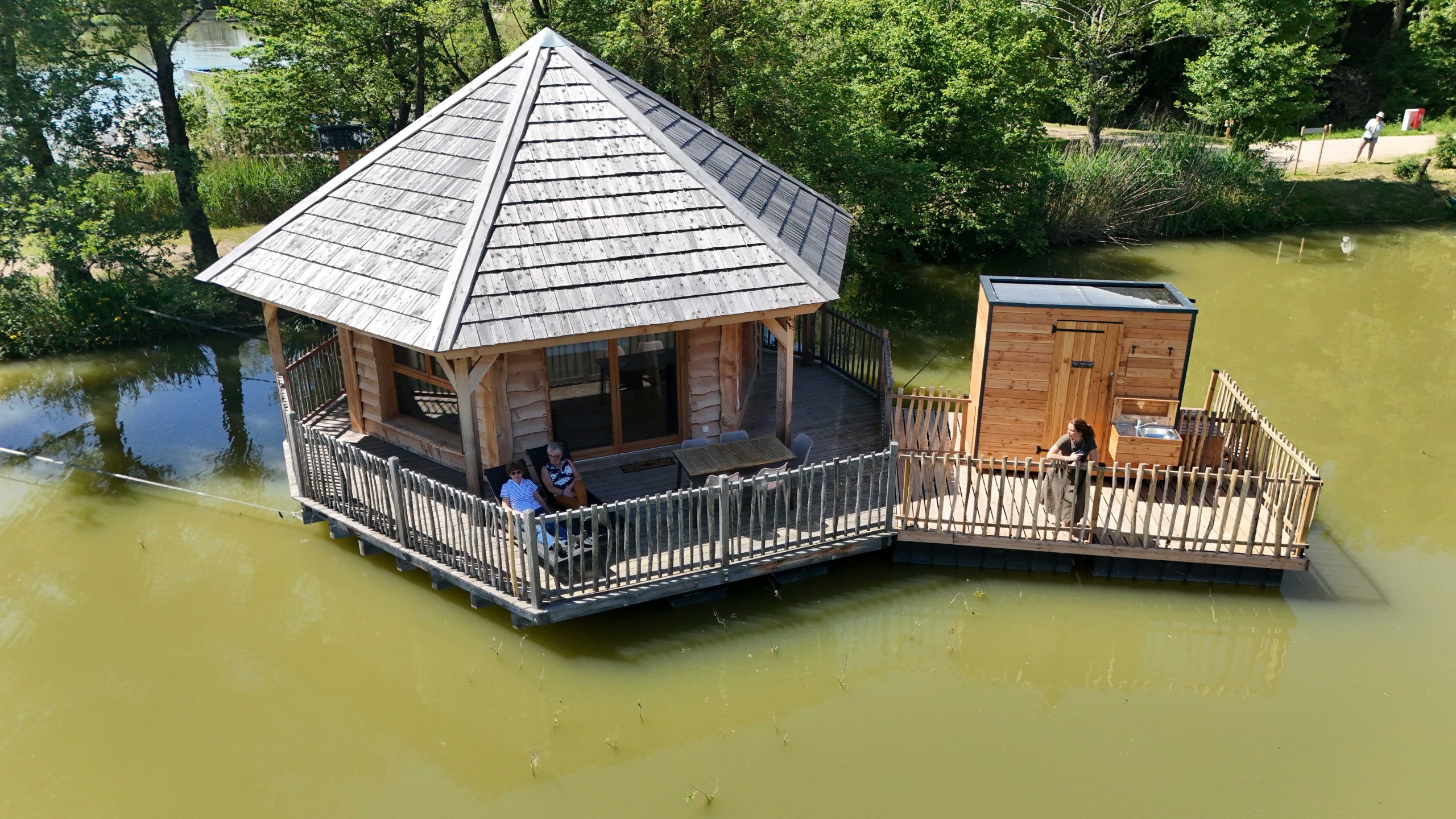 Cabane sur leau avec terrasse en bois, entourée de verdure et dun étang paisible.