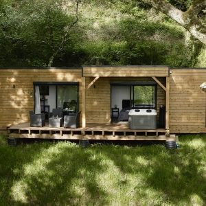 Chalet en bois avec terrasse, entouré de verdure et jacuzzi extérieur.