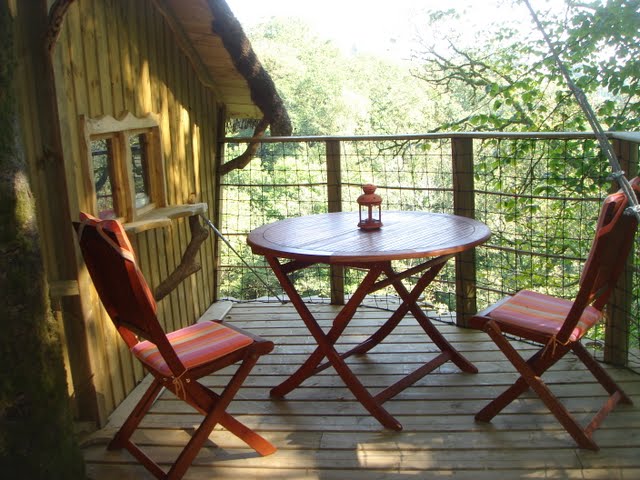 cabanes-dans-les-arbres-bretagne Cabane dans les arbres avec terrasse en bois, table ronde et chaises colorées, vue sur la nature.