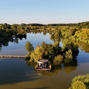 Cabane flottante sur un lac, entourée de verdure et deau calme.
