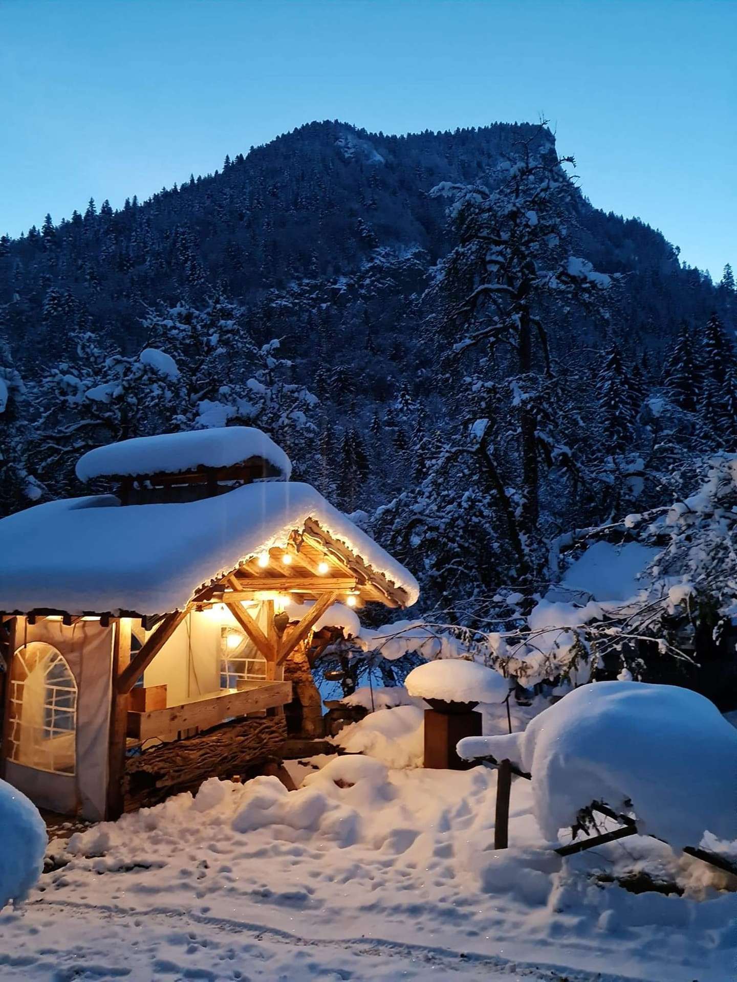 Chalet en bois sous la neige, illuminé par des guirlandes, entouré de montagnes.