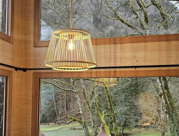 Cabane en bois avec lampes suspendues, vue sur un jardin verdoyant et un petit lac.