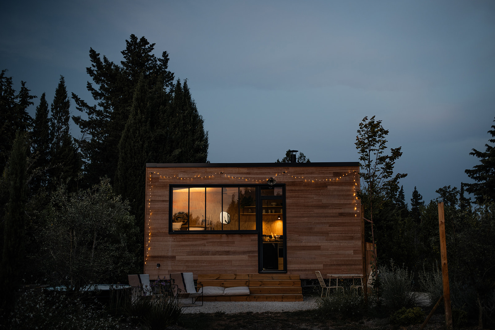 tiny-aout-132 Cabane en bois avec guirlandes lumineuses, entourée d'arbres, créant une ambiance chaleureuse.