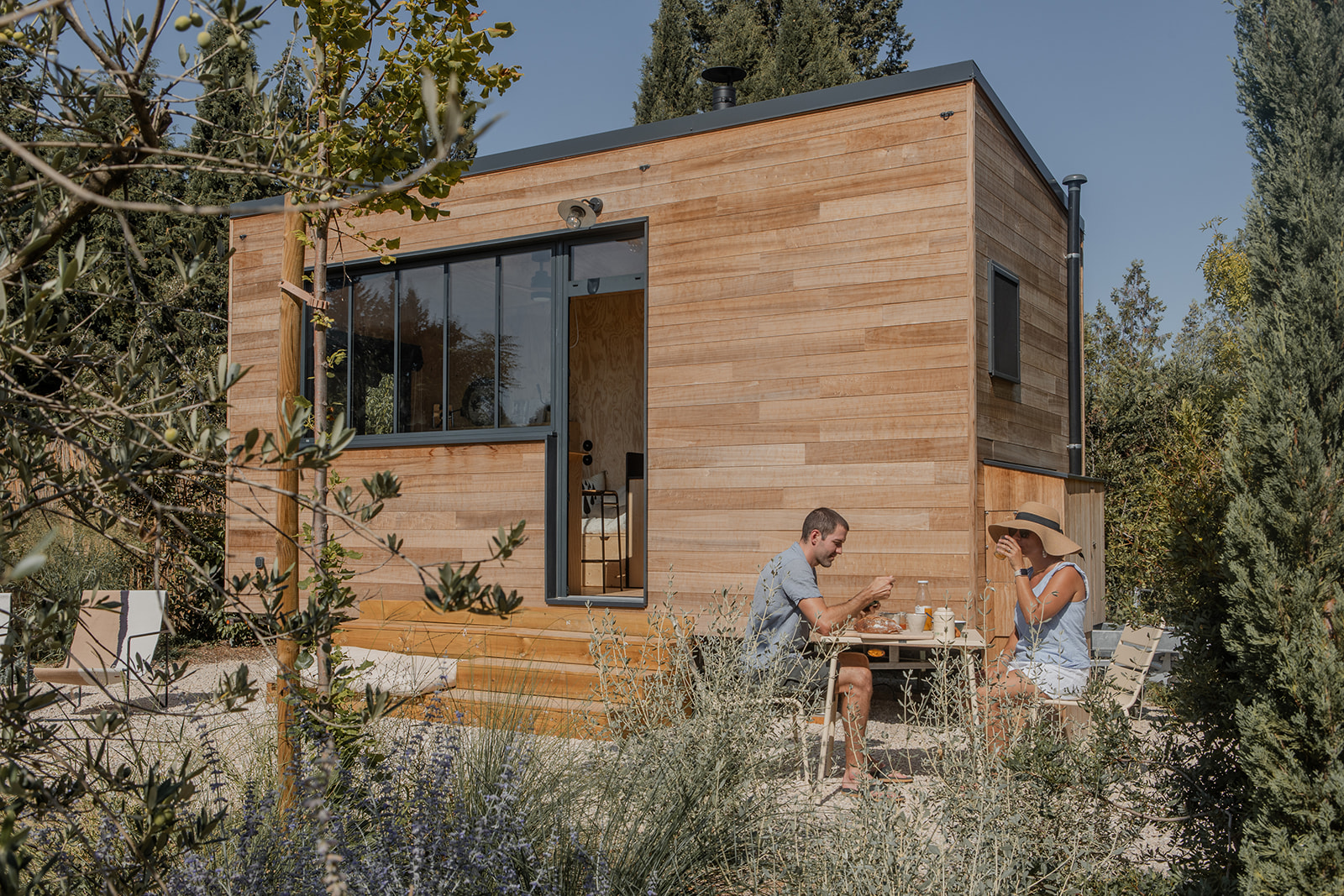 tiny-aout-52 Cabane en bois moderne, entourée de verdure, avec une terrasse et des occupants profitant d'un