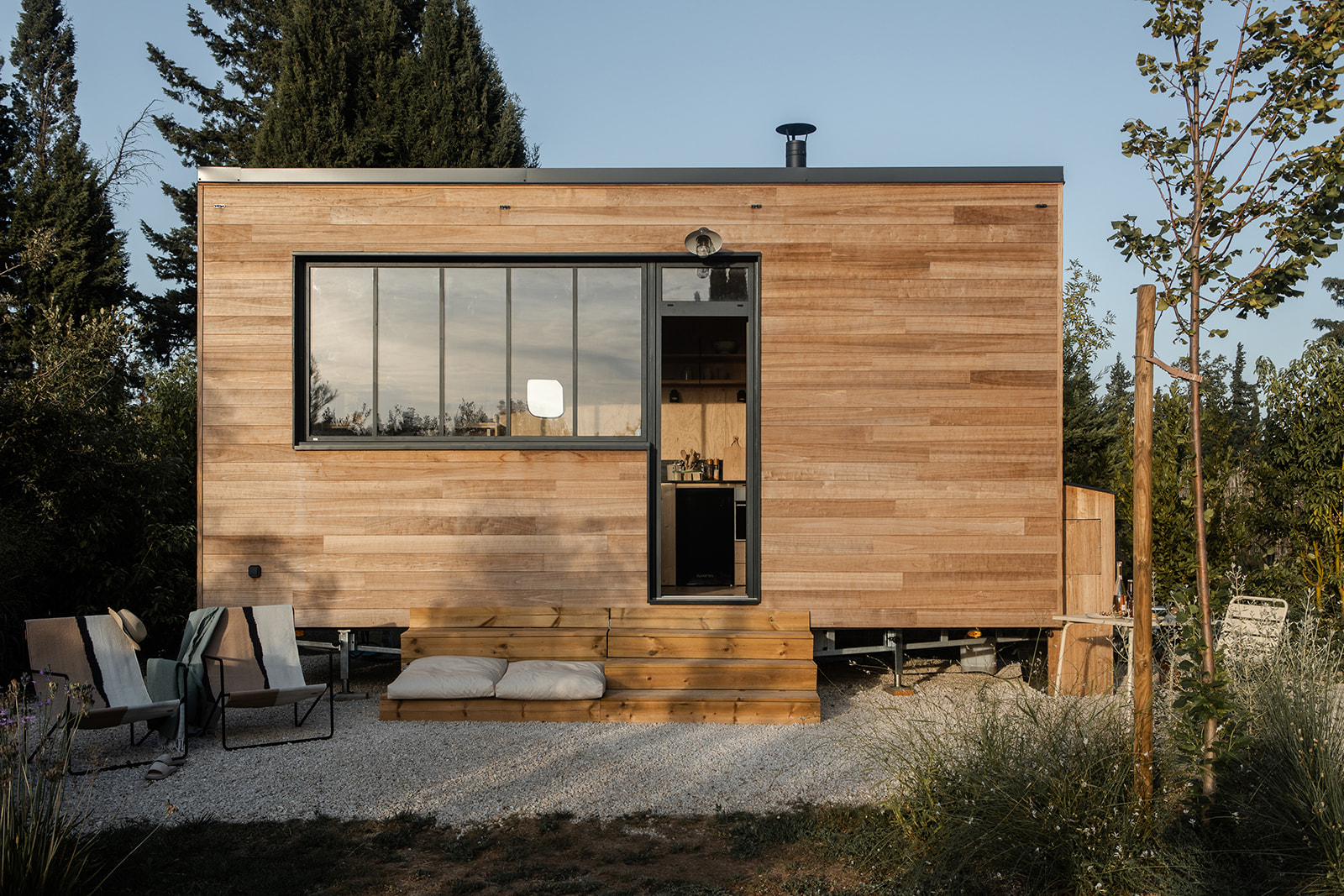 tiny-aout-64 Tiny house en bois avec grandes fenêtres, terrasse en bois et coin détente extérieur.