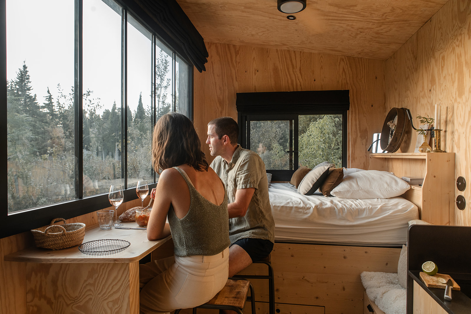 tiny-aout-85 Cabane moderne en bois avec vue panoramique, couple dégustant un repas près de la fenêtre.