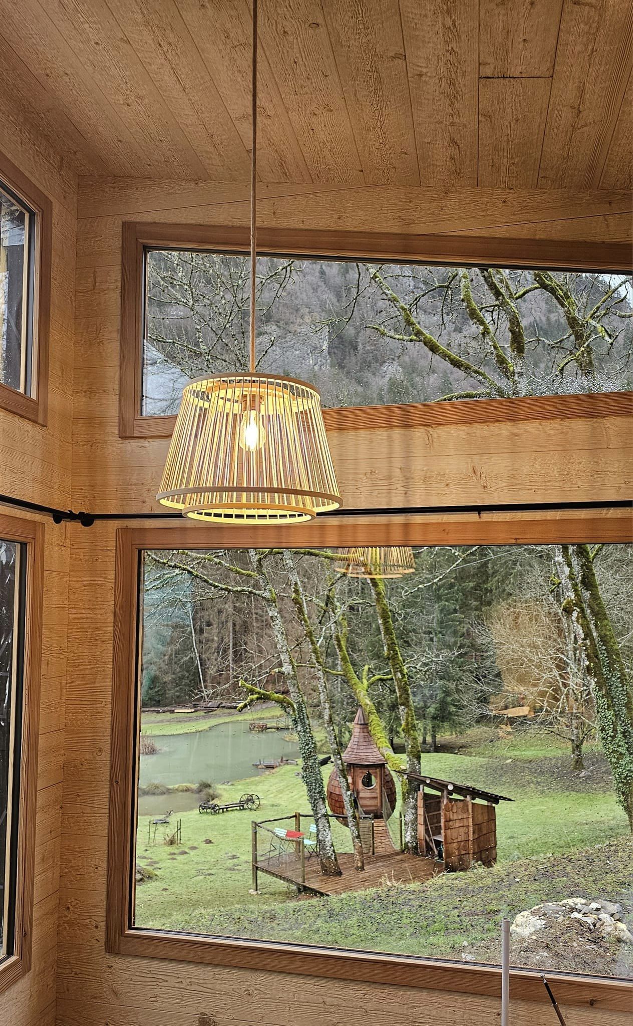 Cabane en bois avec lampes suspendues, vue sur un jardin verdoyant et un petit lac.